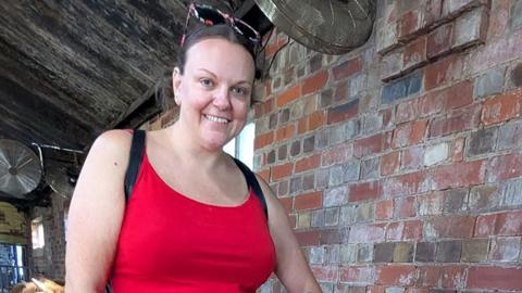 Stacey Waring wearing a red top standing in a barn. She is by herself wearing dark sunglasses on her head and smiling at the camera.