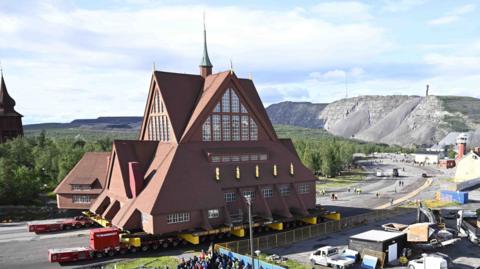 A large brown church dwarfs lorries seen next to it as it's rolled down a road.