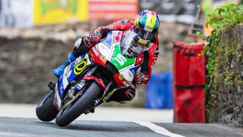 Michael Evans racing on a red, blue and white bike, wearing red leathers and a yellow, blue and pink helmet. A green plate on the front has a white number five on it. There are stone walls and crash barriers on the right and behind him.