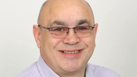 Head image of a man wearing a purple and white striped shirt and glasses, smiling for the camera. 