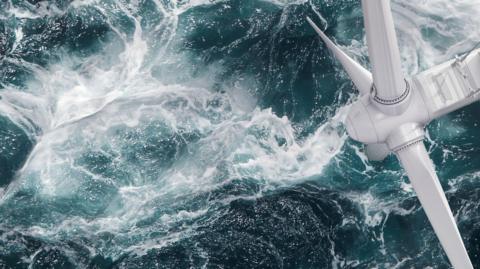 An aerial view of an offshore wind turbine. Visible are the three large white blades and the hub. Below the turbine the sea is green and the water is churned up and there is white froth on the surface of the sea.