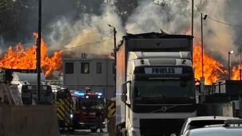 A view of the burning site, with a Fowles vehicle and fire engines. Flames are raging.