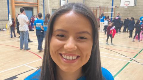 Laura is wearing a blue t-shirt. She has long brown hair and a chain around her neck. She is standing in a busy sports hall with lots of people and children behind her. 