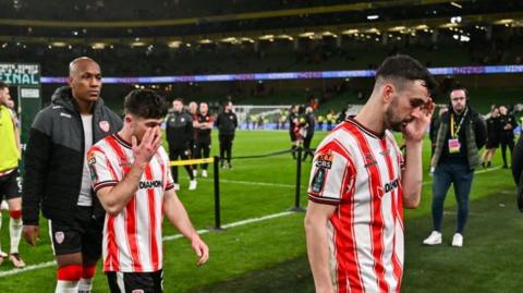 Derry City players in red and white striped football shirt looking down at the ground. 