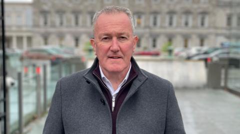 Conor Murphy standing in front of the Irish parliament wearing a grey coat, maroon jumper and white shirt. He is looking a the camera with a neutral expression.