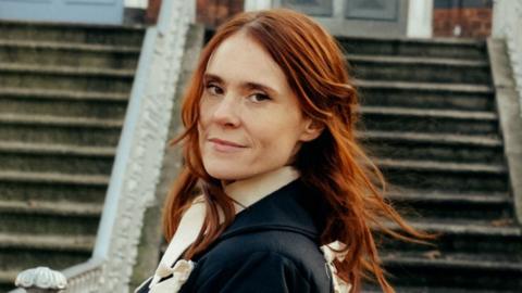 A woman with ginger hair, in a black coat, pictured against some steps