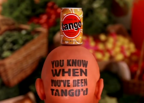 Screen grab showing a back-of-the-head shot of the "Orange Man" character from the iconic 1992 Tango Orange advertisement. A vintage orange Tango can rests on top of his bald, orange-painted head, which is stamped with the classic brand slogan: "You Know When You've Been Tango'd"