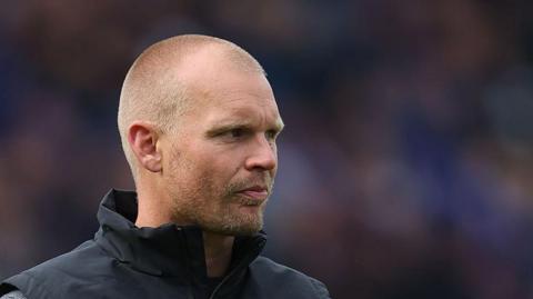 Norwich City manager Liam Manning looking thoughtful to his left, wearing a sleeveless rain jacket 