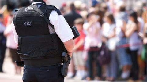 A police officer stands watching a crowd, a small gun holstered at their hip