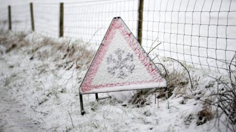 A triangular road sign warning of snow and ice, partially covered in snow. The grass around the sign is covered in snow, with a wire fence and a snowy field in the background.