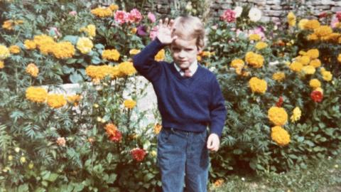 A grainy film photograph from the 1980s showing a young boy, perhaps aged about four, waving as he stands in a garden. He is wearing a white shirt, red tie, navy jumper and blue cords. Behind him are lots and lots of flowers, mostly yellow although there are red, orange and pink flowers too, and a Cotswold stone wall.