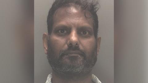 Police custody image of Nimalarajah Mathiyadaranam with black hair and beard wearing a white patterned shirt. 
