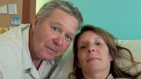 A man and woman in a selfie image. She is propped up in a hospital bed and has long brown hair. He is leaning his head towards hers and has grey hair and a beige polo shirt. Neither are smiling.