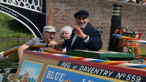 Sir David Suchet with dark hair and beard wearing a dark-coloured cap and rdark-coloured jacket is at the stern of a narrowboat moving the tilled with one hand and waving with the other. There is a man in a light brown cap and a woman with white hair behind him.  There is a painted water can on the roof of the boat alongside a chimney. The boat has colourful paintwork and the words REG AT DAVENTRY in white lettering on the side. The boat has just passed through an iron bridge painted in black and white.