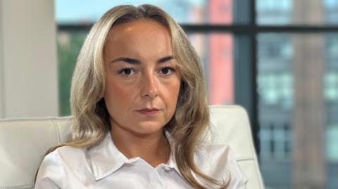 Chloe Gibson is looking at the camera with a stern expression on her face. She is wearing a white shirt and has long blonde hair.