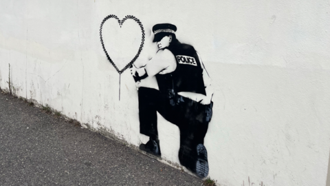 A spray painted stencil of a police officer kneeling down with a spray paint can which has drawn a love heart 