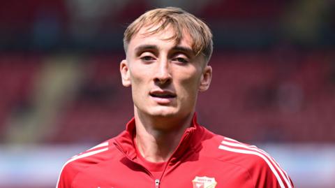 Jake Tabor wearing a red Swindon Town jacket and standing on the pitch during a pre-game warm-up in August 2025.
