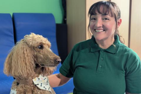 Katia Stanford, a woman with dark hair tied back and a fringe, wearing a dark green polo shirt, smiles at the camera with her hand on a biscuit-coloured poodle.