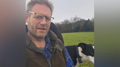Steven Conisbee is wearing a brown gilet over a blue shirt with his black and white dog in the background in a selfie photograph. There are fields and woods behind him.