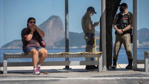 A woman wearig a face mask waits for a bus next to municipal guards, during the first day of lockdown due to the COVID-19 coronavirus, at Icarai neighbourhood in Niteroi, Rio de Janeiro state, Brazil, on May 11, 2020.