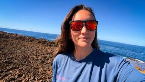 Lara Vafiadis takes a selfie while wearing orange tinted sunglasses and a blue top. She has long brown hair and is standing in front of the ocean on a sunny and clear day.
