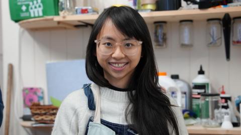 Cecilia is wearing clear rimmed glasses and wearing her long, black hair and down over her left side. She's wearing dark blue denim dungarees and a pale blue apron with a white jumper. She's facing the camera and smiling.