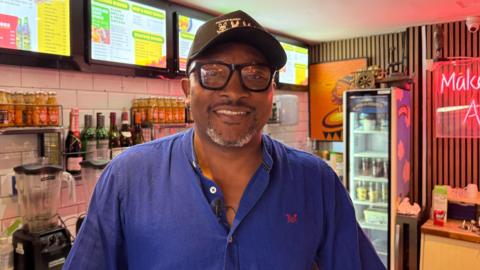 A man smiling and wearing a black hat with black glasses. He has a blue shirt on with a red logo. 
