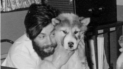 A man with dark hair and a dark beard holding a husky dog