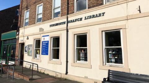 The cream slightly faded exterior of Tynemouth Branch Library. Books can still be seen on shelves through the windows. A bench has been placed outside near the bottom of an accessibility ramp to the front door.