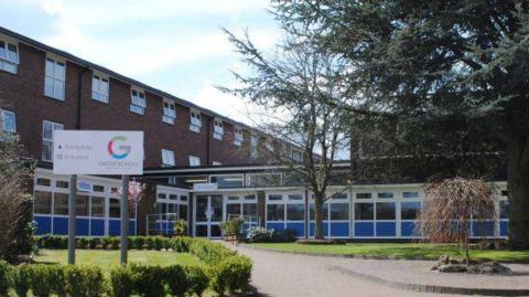 A school building seen from outside. A sign can be seen for Grove School in a grassy landscaped area. It is a three story building. There is blue cladding below windows on the bottom floor.