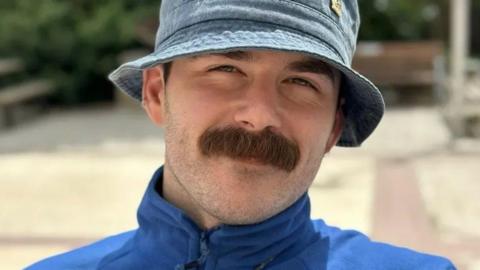 A man with a moustache, wearing a blue zip up jumper and denim hat. He is looking directly at the camera. Behind him the background is blurred. There is concrete, a bench and bushes. 