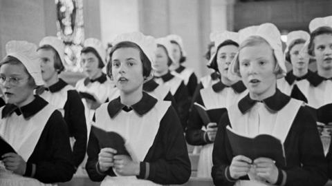 A black and white image showing young girls in old fashioned pinafores and white hats, singing at a chapel service held at the Foundling Hospital in Berkhamsted in the spring of 1941