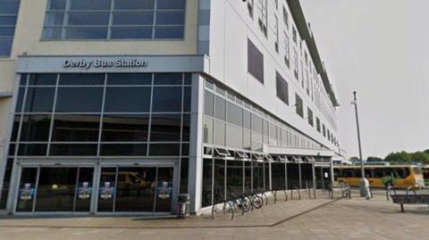 A large grey building that is mostly windows on the ground floor. A yellow bus can be seen to the left.