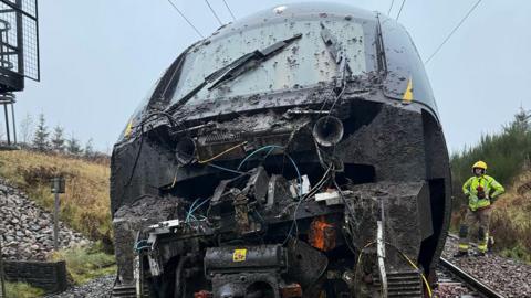The front of the train which looks badly damaged and is covered in mud. There are wires visible. There is a man in hi-vis standing to the right with his hands on his hips.