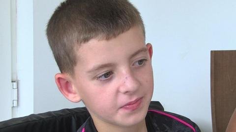 Bradley sitting, looking to the side, wearing  a black and pink t-shirt, in front of a  plain light-blue wall