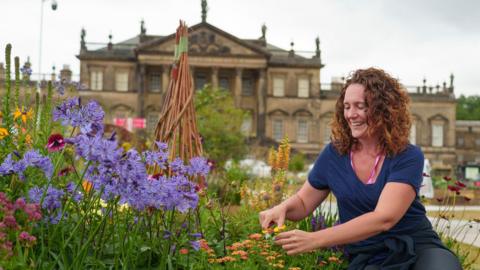 A woman with curly auburn hair and wearing a blue t shirt smiles as she trims back plants in an organised flowerbed with many types and colours of flowers. Wentworth Woodhouse, a stately home, is blurred in the background.
