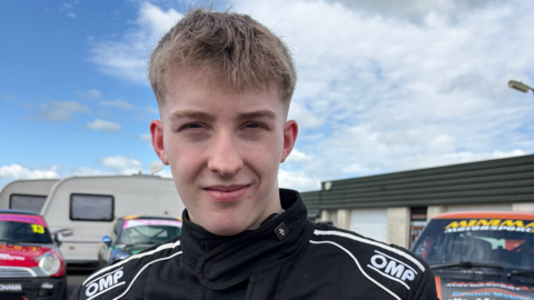 Ally Chalmers, a 17-year-old boy with shot, blond hair. He is looking at the camera and is half smiling. He is wearing a black race suit with the names of sponsors on it. There are racing cars behind him.
