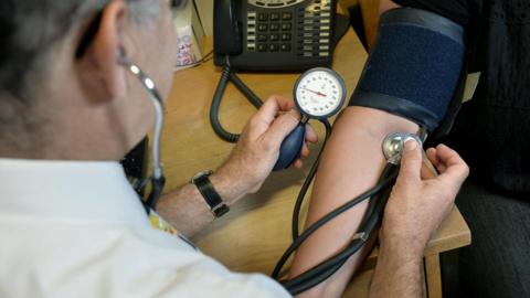 A doctor conducting a blood pressure test on a patient. He is holding a medical instrument against an arm whilst monitoring a gauge. The patient has a dark blue wrap around their arm.