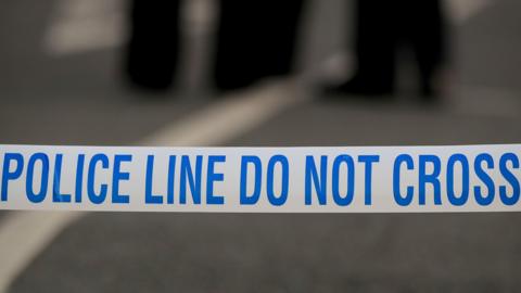 Stock image of police tape which says "Do not cross" in blue on white tape. The background is blurry.