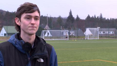 Joseph Russell, Sgioba Loch Aillse, na sheasamh aig oir na pàirce agus seacaid dubh is ghorm air. 