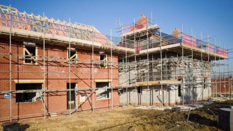 Homes under construction as part of a new-build housing development. The partially-built houses have scaffolding around them and the roofs are not yet finished. There is a clear blue sky.