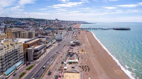 An aerial view over Brighton and its seafront. People are on the shingle beach and the pier is in the background. On the left there is the city.