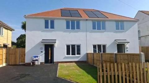 A whit semi-detached house with a driveway to the left. small lawn to the right and wooden fence in the foreground.