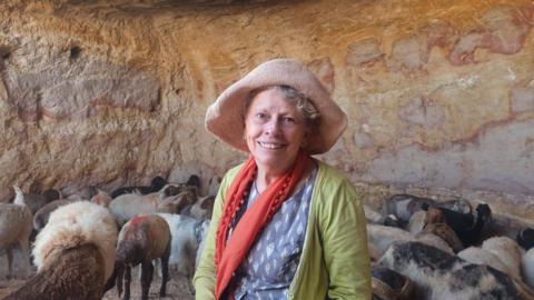 A woman smiling at the camera wearing a hat, red scarf, green jumper and a blue top. You can see a group of sheep behind her and she appears to be in a .cave