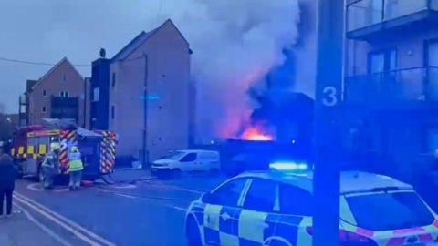 Fire crews and a fire engine can be seen in a street. Flames and smoke can be seen near flats and a police car is in the foreground. 