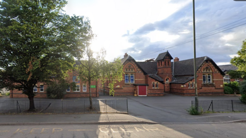 The front of Holmer Academy. It is a red brick school building with a grey roof and large concrete area for cars and a black metal fence. There is a road outside the fence.