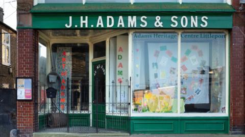 A green painted shop with large plate glass windows and a gated covered foyer to its entrance, with tiled flooring. 
