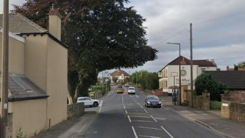 A road stretches into the distance. On the left is a cream-coloured building, a tree, and the front of a white car poking out of a side road. On the right is a low road, a pub in the distance with the words The Royal written on the side of the building.