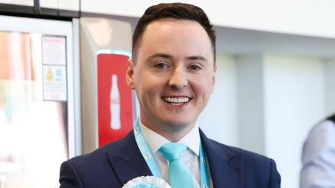 Darren Grimes is smiling at the camera. He is wearing a navy suit with a light blue tie and a Reform rosette in the same colour. His short brown hair is swept to the side. 