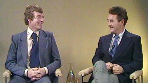 Two football pundits wearing suits sit in chairs smiling during studio debate.